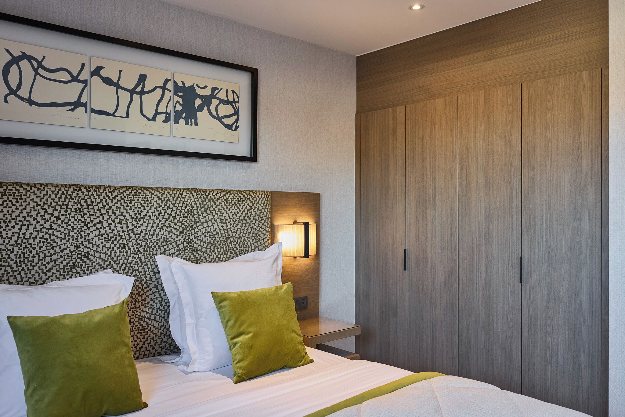 A hotel room with a bed, pillows, a headboard with a patterned design, a wall-mounted artwork, and a wardrobe.