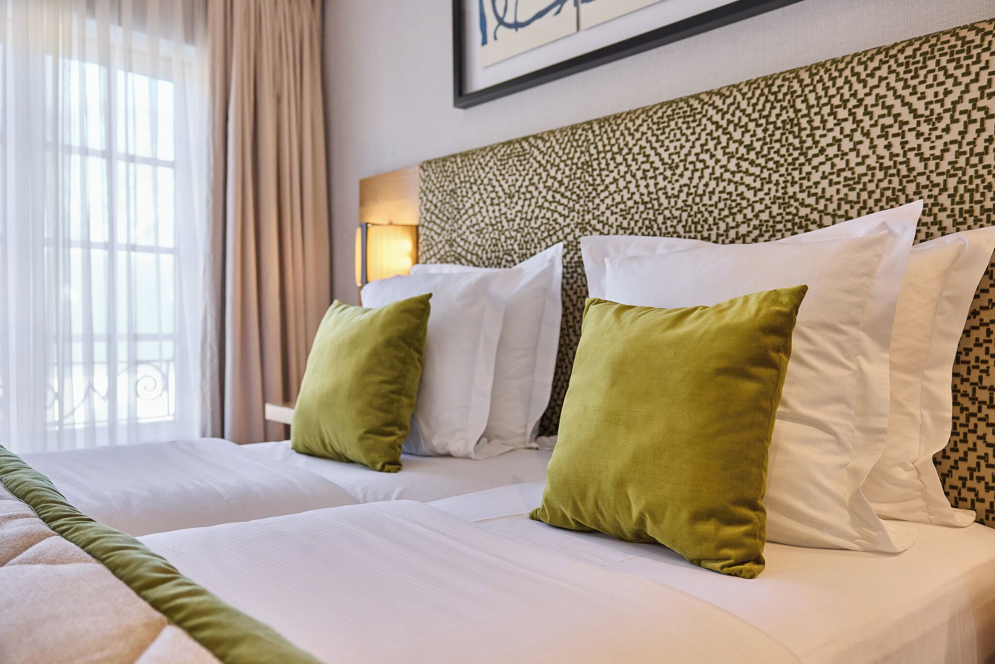 A hotel room with two beds, each with white linens and green pillows. The room has a patterned headboard and a window with curtains.