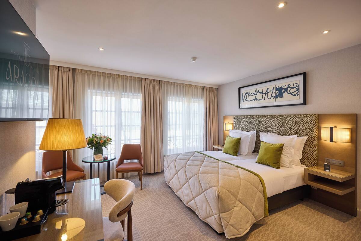 A hotel room with a bed, bedside tables, a desk with a lamp, chairs, and a view of the outdoors through large windows.