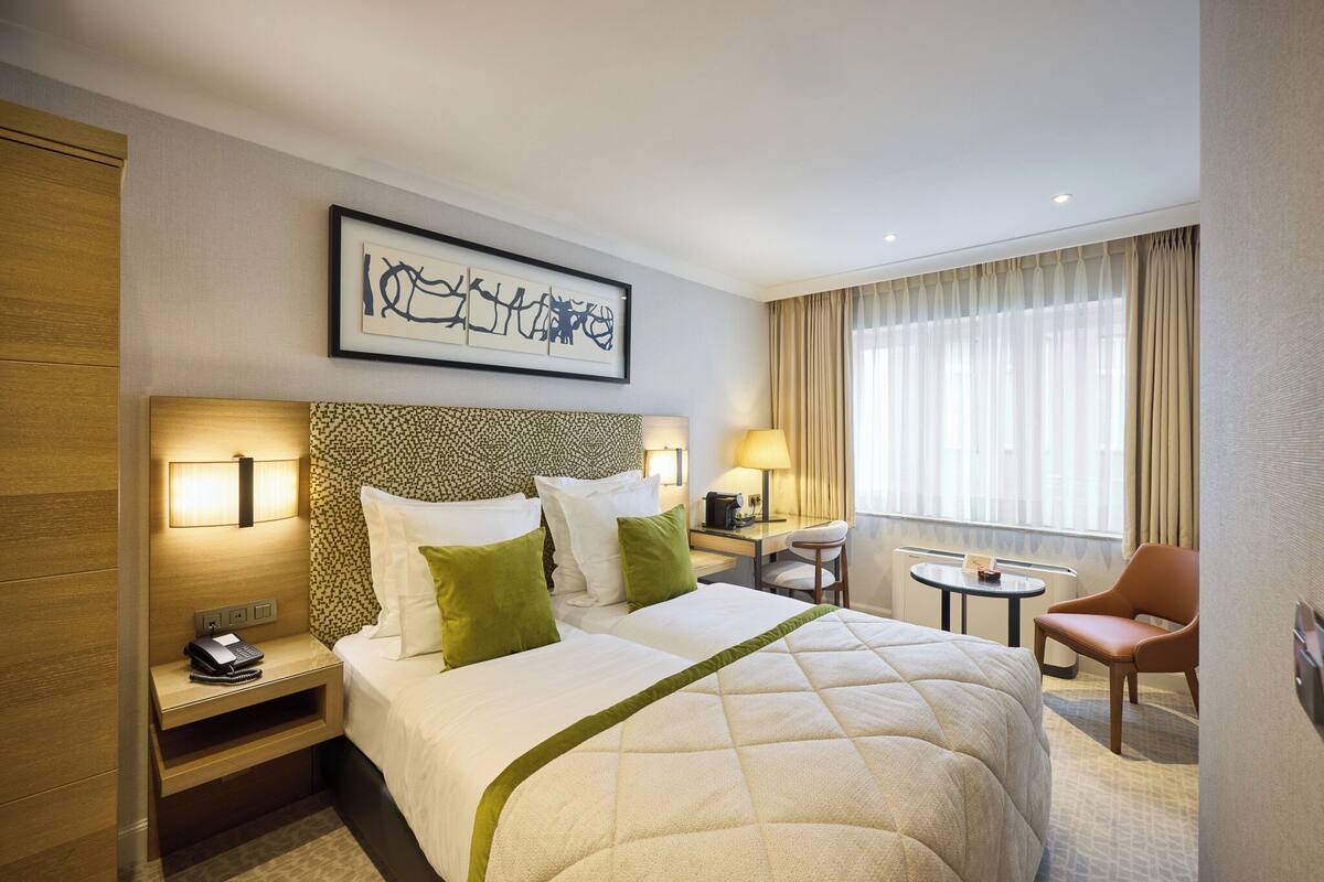 A hotel room with a large bed, bedside tables, a desk, a chair, and a window with curtains.