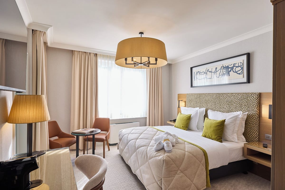 A hotel room with a bed, a desk, a chair, a lamp, and a painting on the wall.