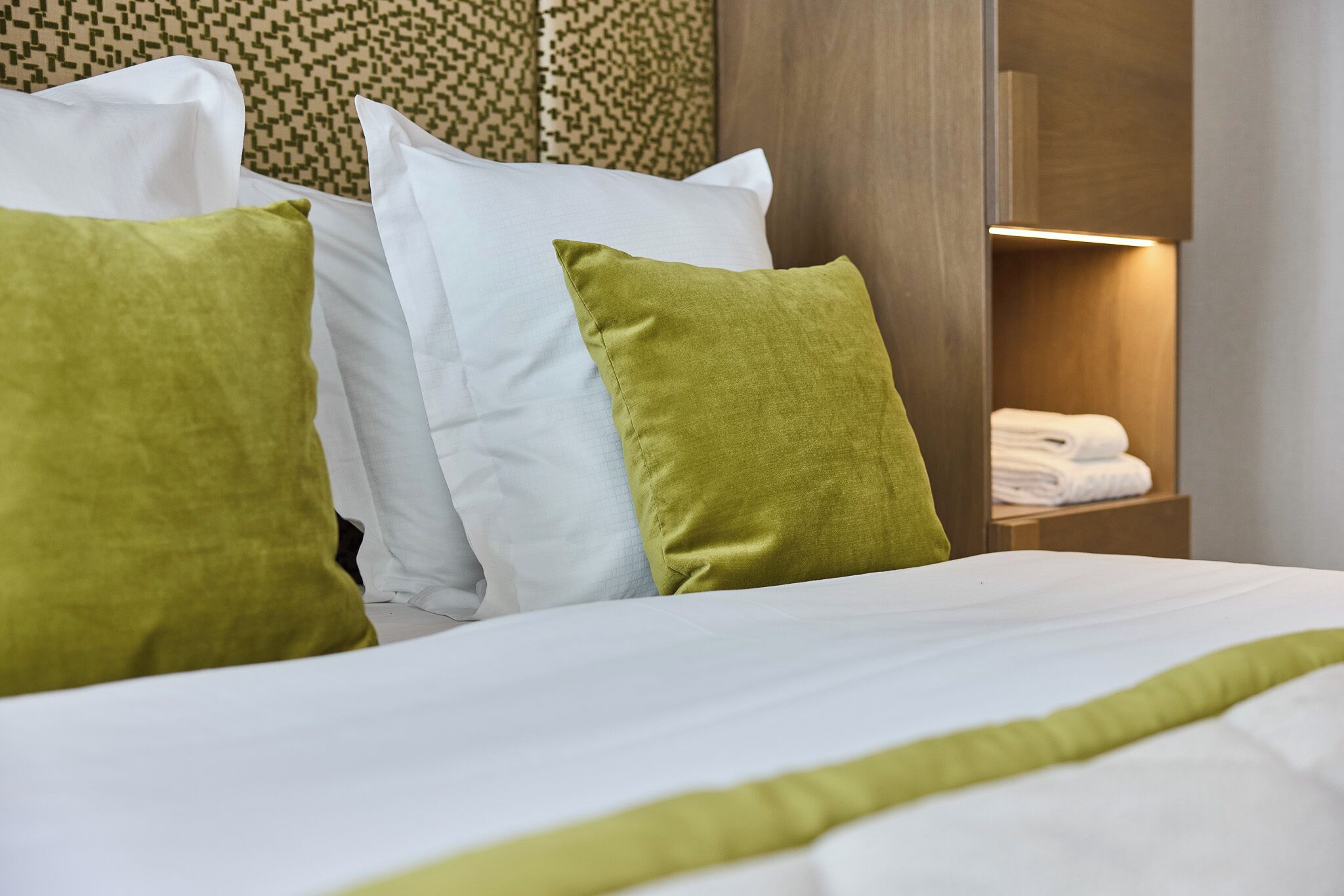 A neatly made bed with white and green pillows, a wooden headboard, and a small wooden shelf with folded towels.