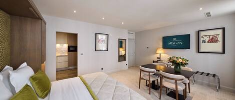 A hotel room with a bed, a desk with a chair, a small table with a vase of flowers, and framed pictures on the walls.