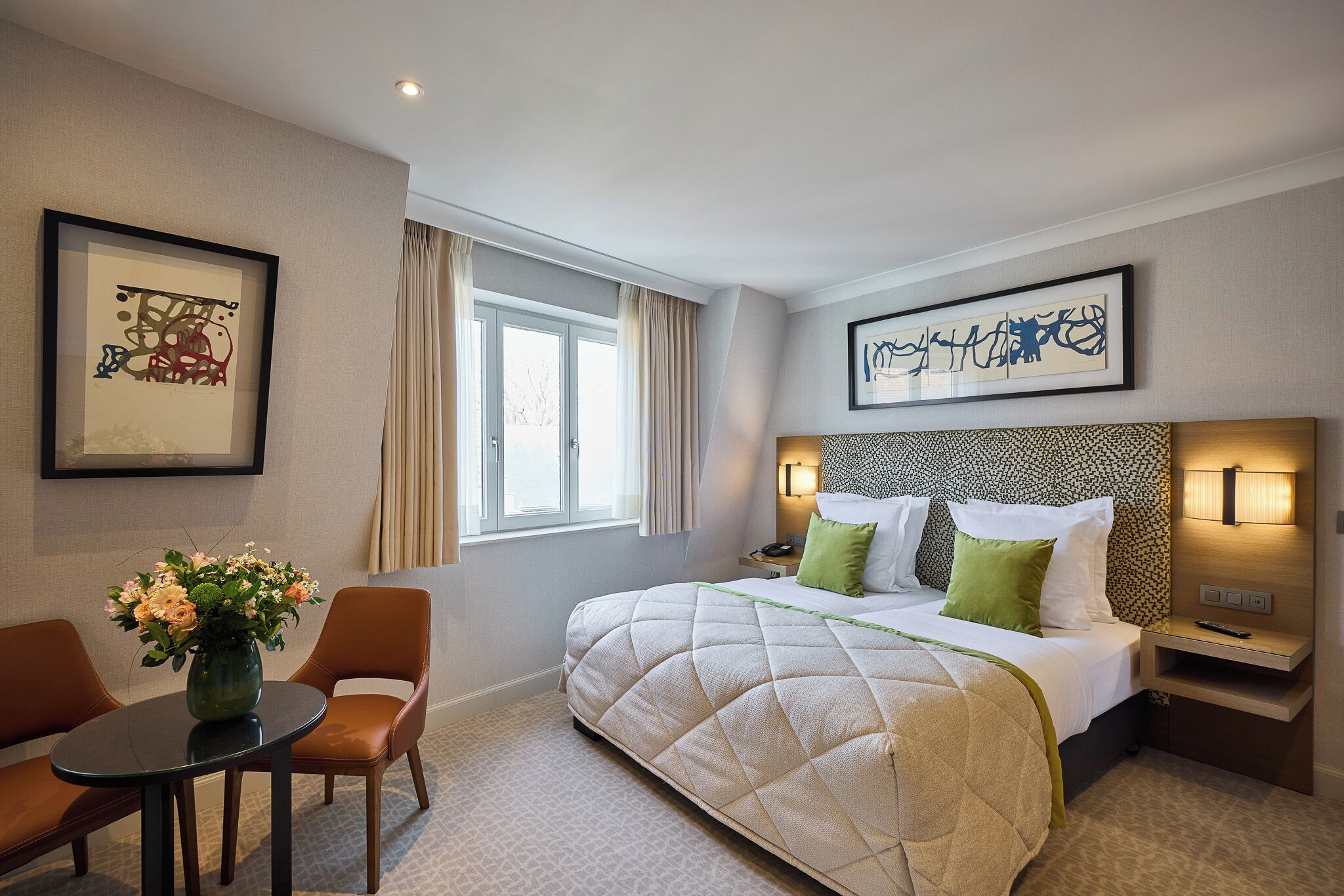 A hotel room with a large bed, a bedside table with a lamp, a small round table with chairs, and artwork on the walls.