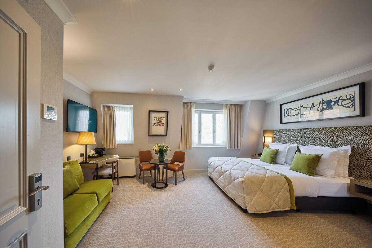 A hotel room with a bed, a green sofa, a small table with chairs, a desk, a lamp, and a painting on the wall.