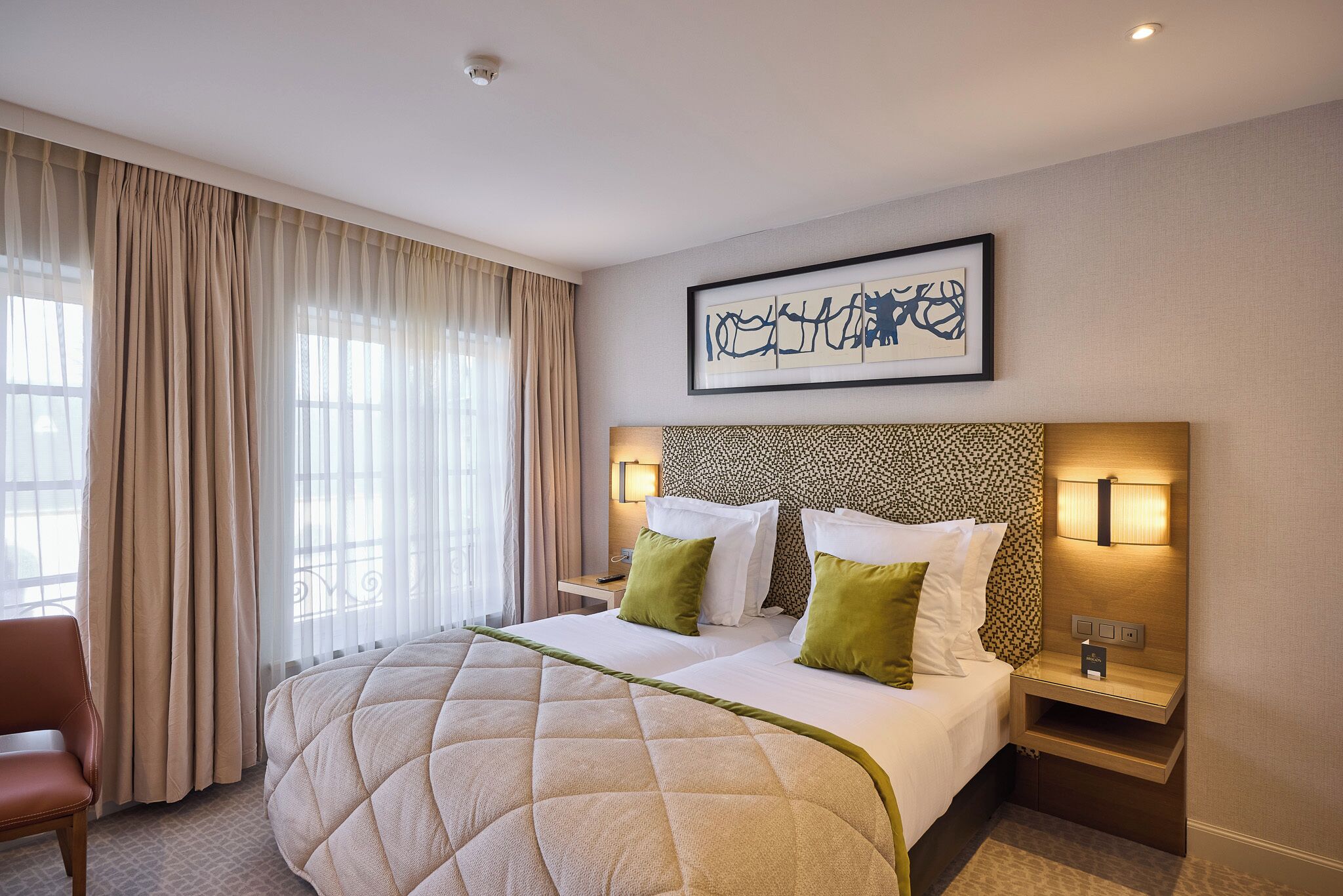 Quarto de hotel com uma cama grande, duas mesas de cabeceira, uma cadeira e uma janela ampla com cortinas.