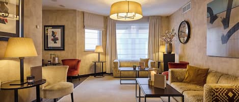 A hotel room with a sofa, armchairs, a coffee table, a lamp, a clock, and a window with curtains.