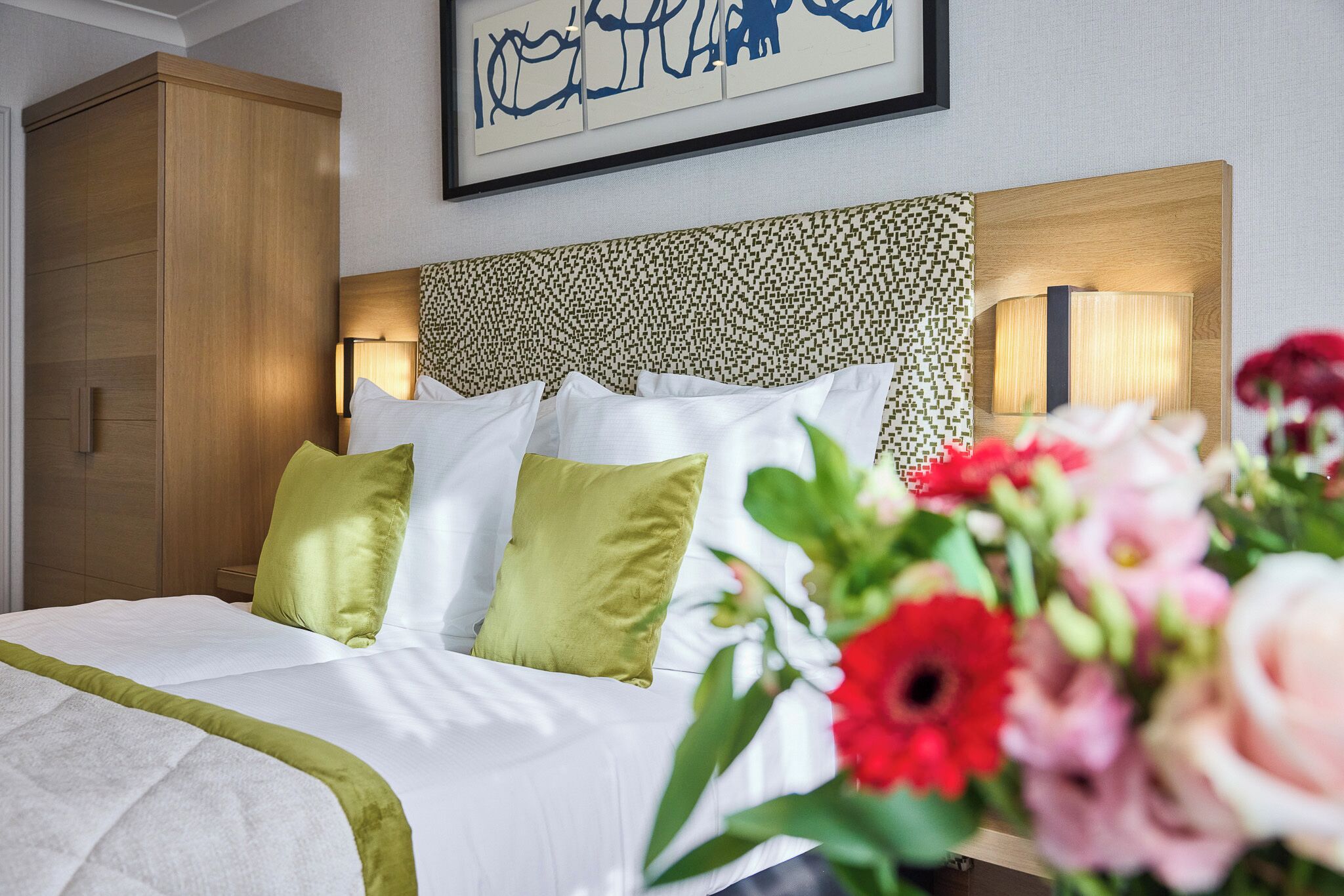 Une chambre d’hôtel avec un lit, une tête de lit ornée d’un motif décoratif, une lampe de chevet et une œuvre d’art encadrée au-dessus du lit.