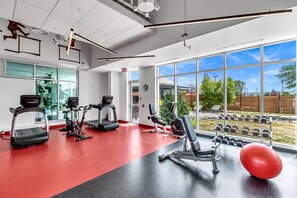 Une salle de sport équipée de tapis de course, de vélos elliptiques et de poids libres, avec de grandes fenêtres et un sol rouge.