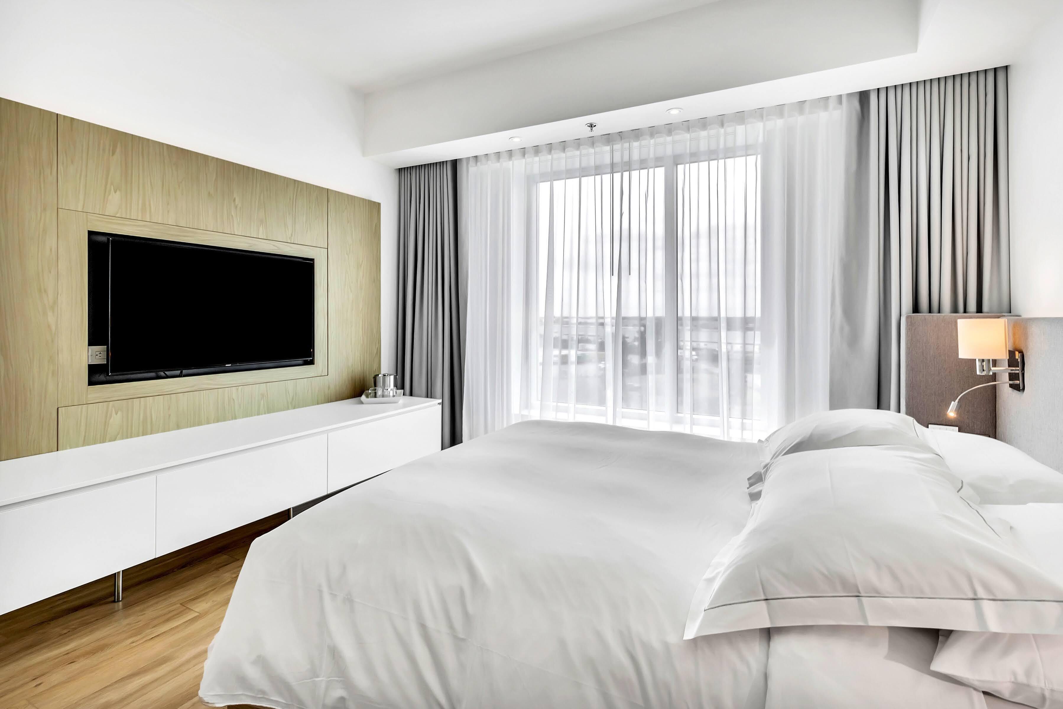 A modern hotel room with a large bed, a flat-screen TV mounted on a wooden panel, a bedside table with a lamp, and a large window with sheer curtains.