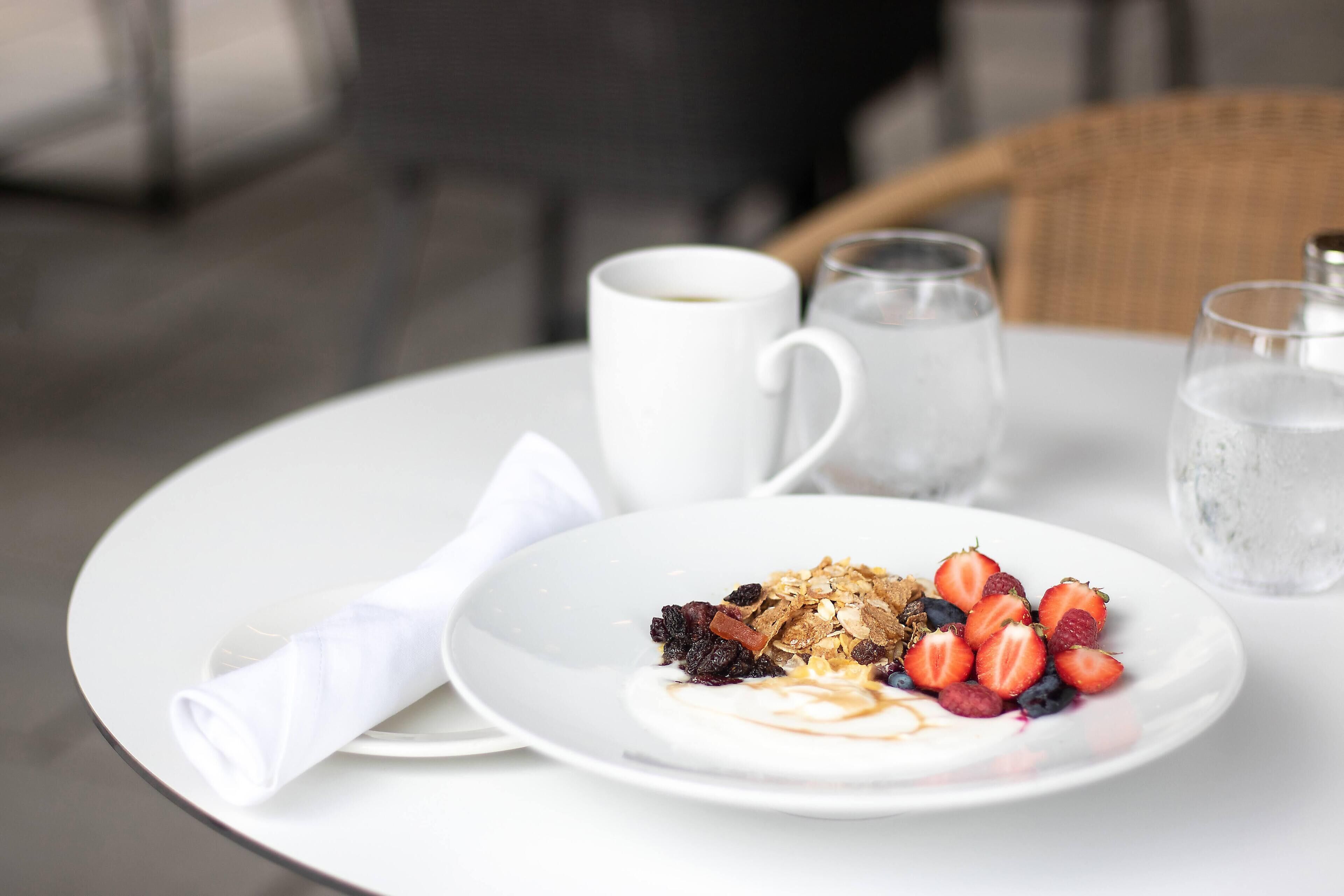 Un plato blanco con un desayuno de yogur, granola, fresas y arándanos sobre una mesa blanca.