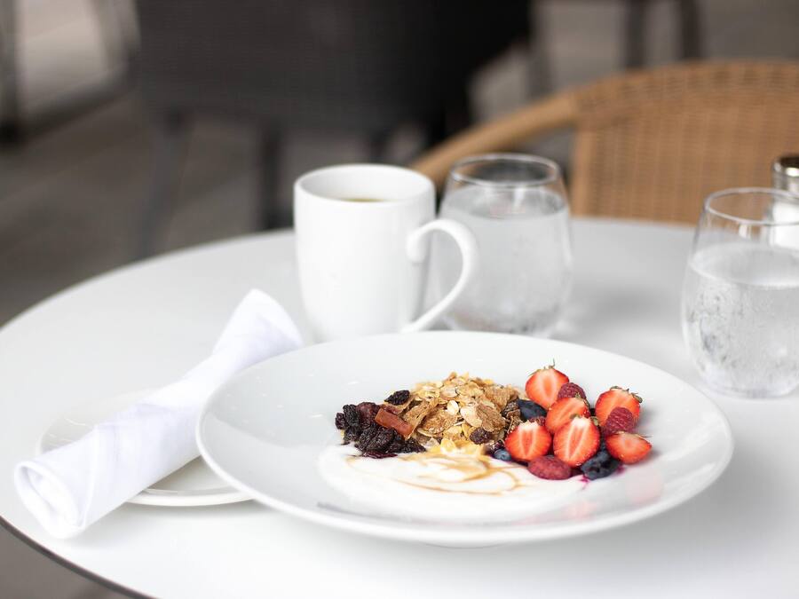 Un plato blanco con un desayuno de yogur, granola, fresas y arándanos sobre una mesa blanca.