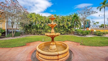 A circular stone fountain with a tiered design, surrounded by a brick pathway and lush greenery.