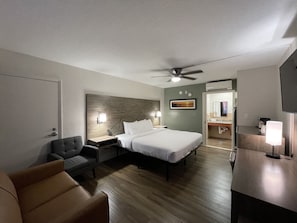 A hotel room with a bed, a desk, a chair, a ceiling fan, and a bathroom visible through an open door.
