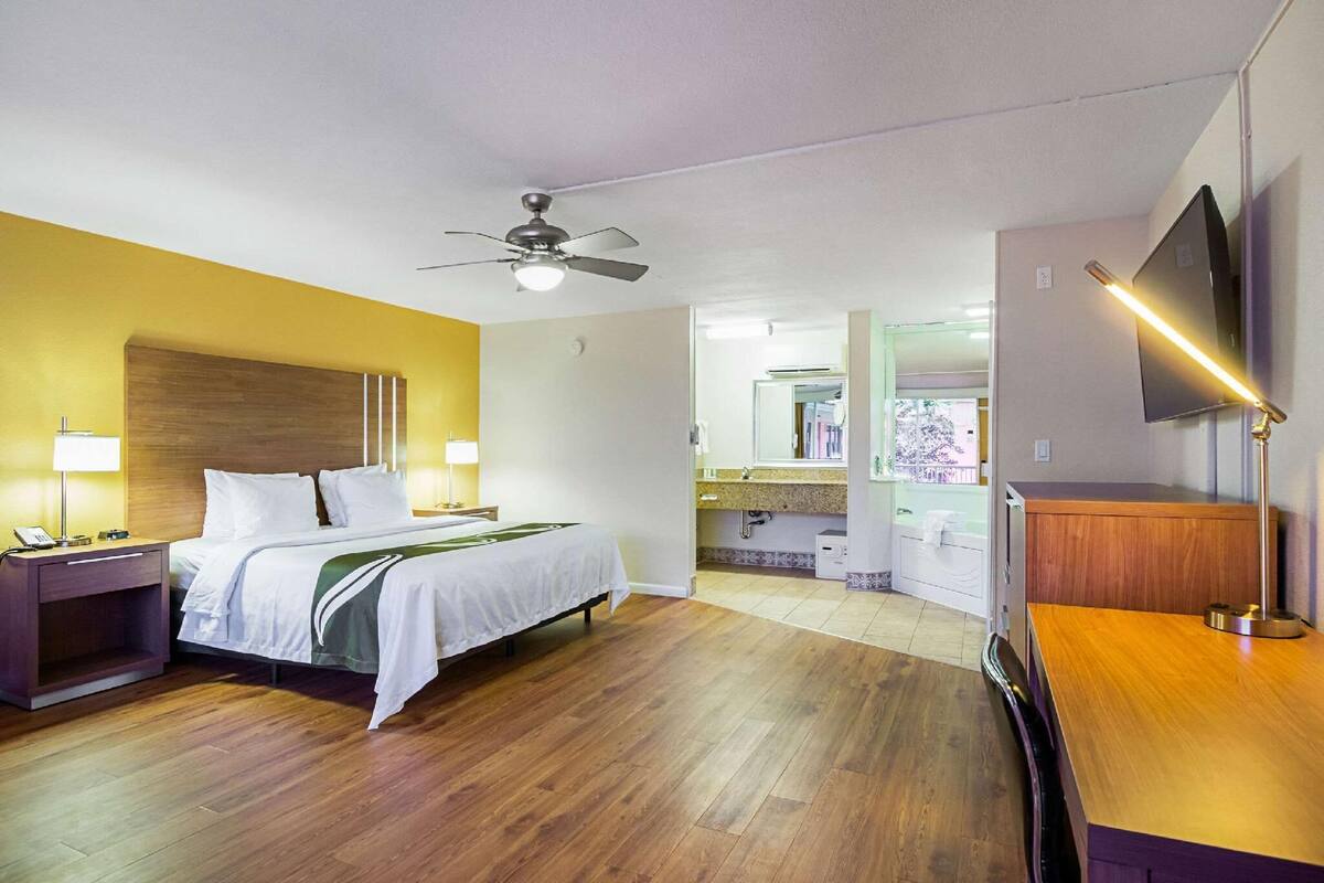 Hotelkamer met een bed, nachtkastjes, een plafondventilator, een televisie en een badkamer zichtbaar door een open deur.