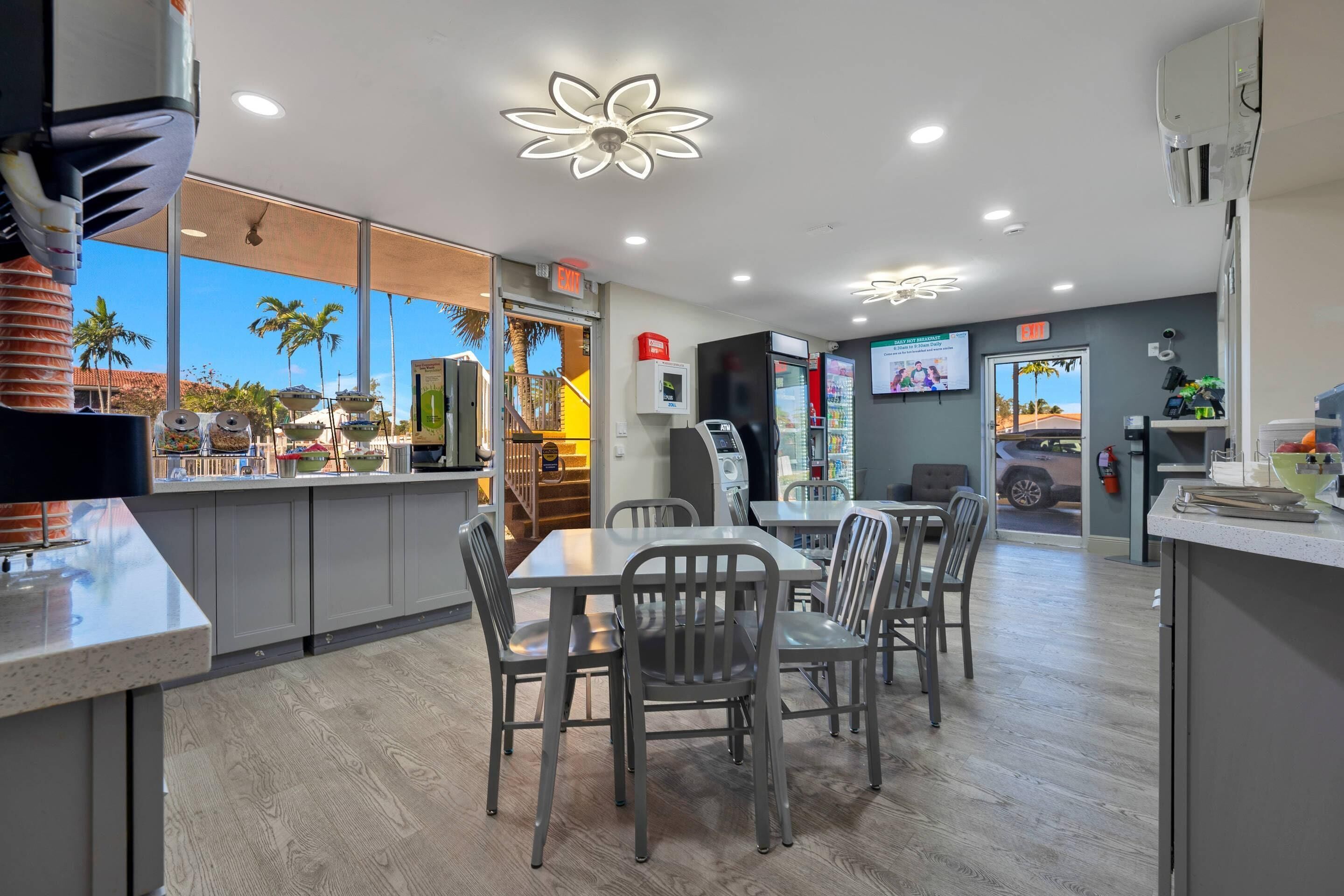 Interior moderno de una cafetería con comedor, cocina y vistas a palmeras.
