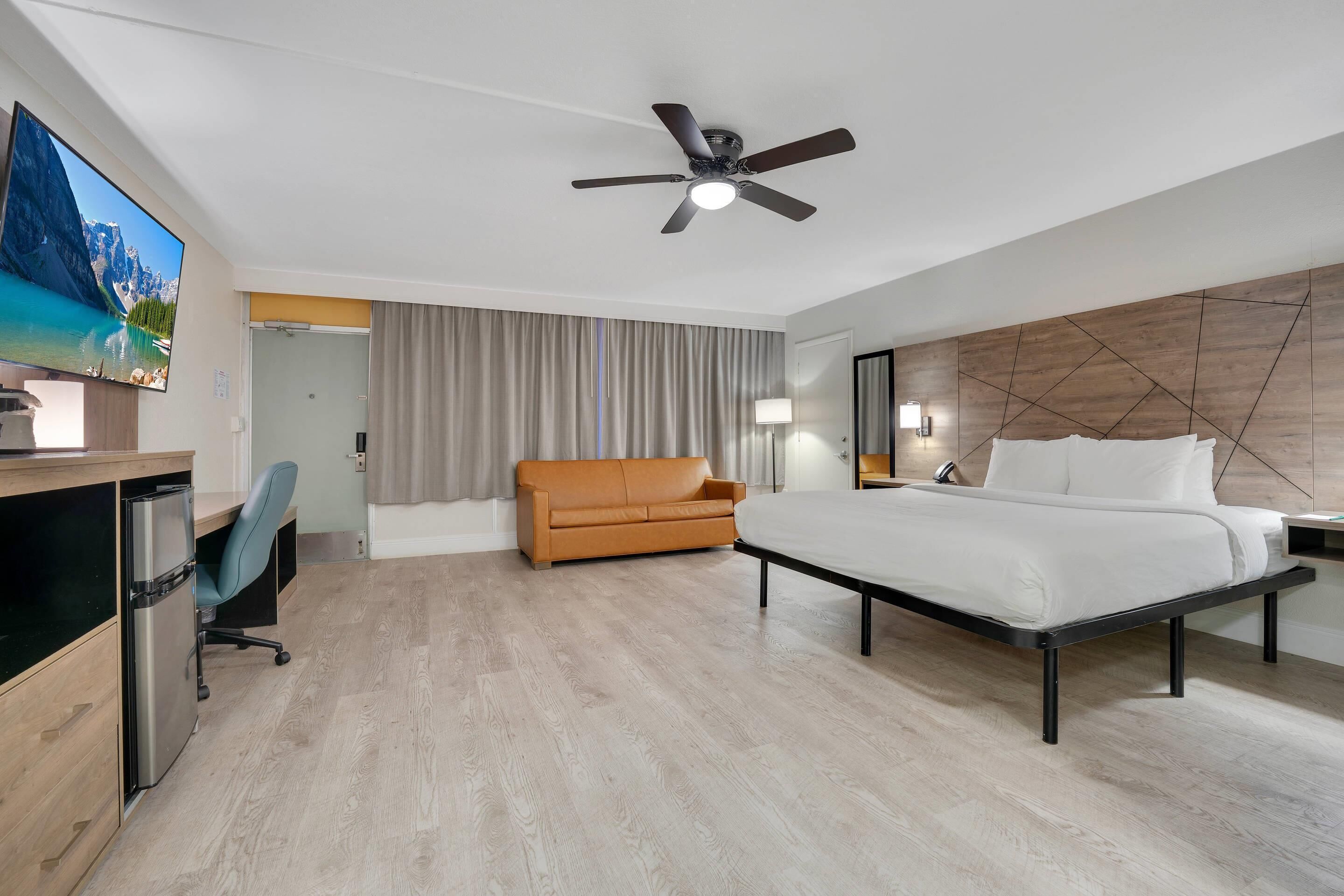 A hotel room with a bed, a desk with a computer, a television, a ceiling fan, and a view of a mountain lake.