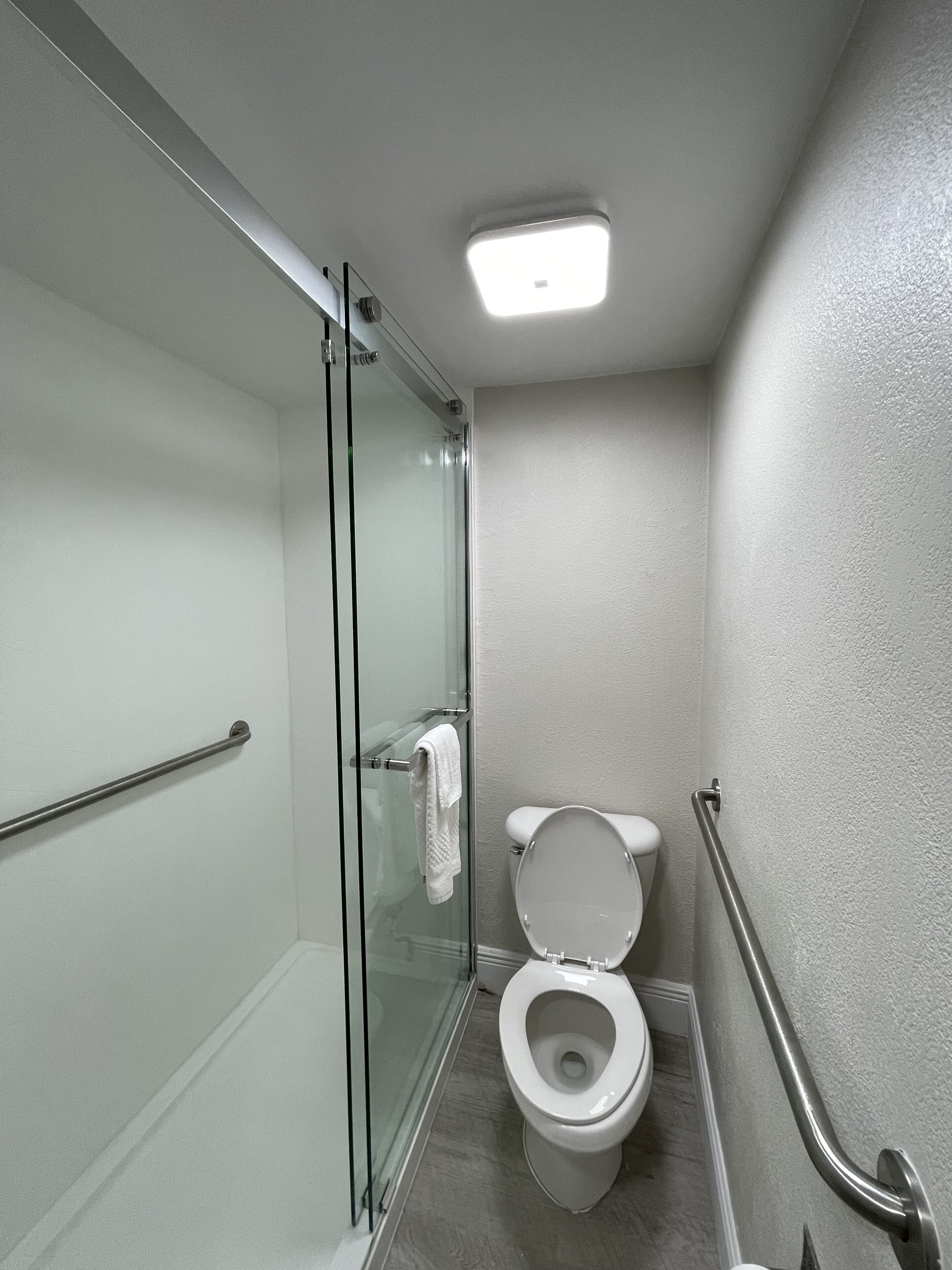 Casa de banho pequena e moderna, com cabine de duche em vidro, sanita branca e barra de apoio.