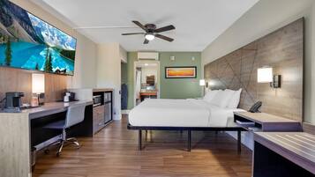 Une chambre d’hôtel avec un lit, un bureau et une chaise, un ventilateur de plafond, un tableau et une vue sur un lac de montagne.