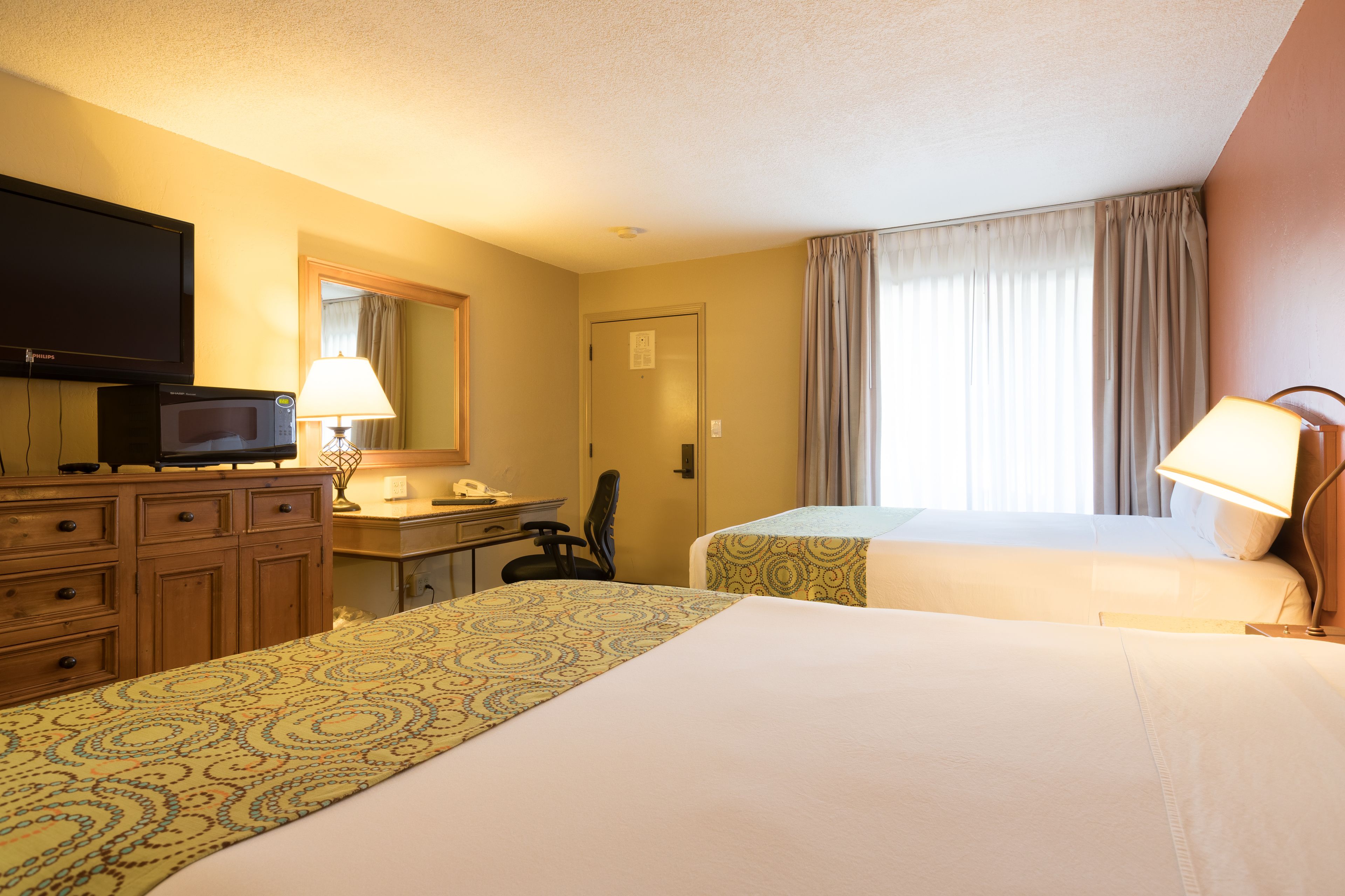 A hotel room with two beds, a desk with a lamp, a television, and a window with curtains.