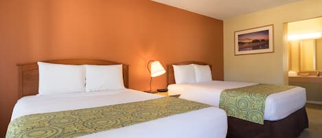 A hotel room with two beds, each with white linens and a patterned throw. There is a nightstand with a lamp between the beds, and a framed picture on the wall.