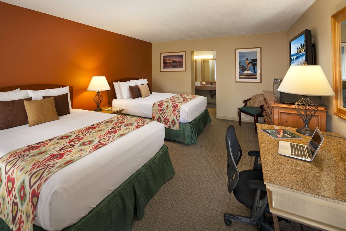 A hotel room with two beds, a desk with a laptop, a chair, a desk lamp, a television, and artwork on the walls.