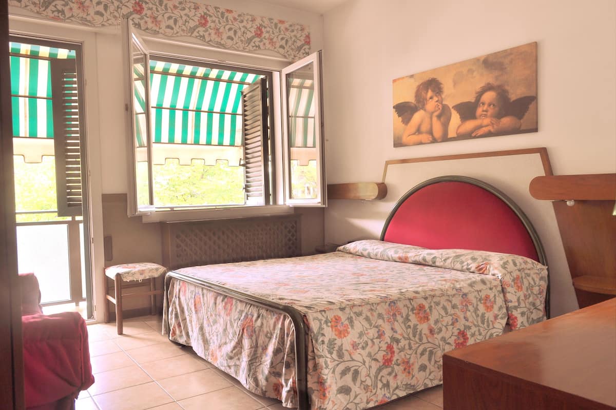 A bedroom with a bed, a chair, a window with shutters, and a painting on the wall.