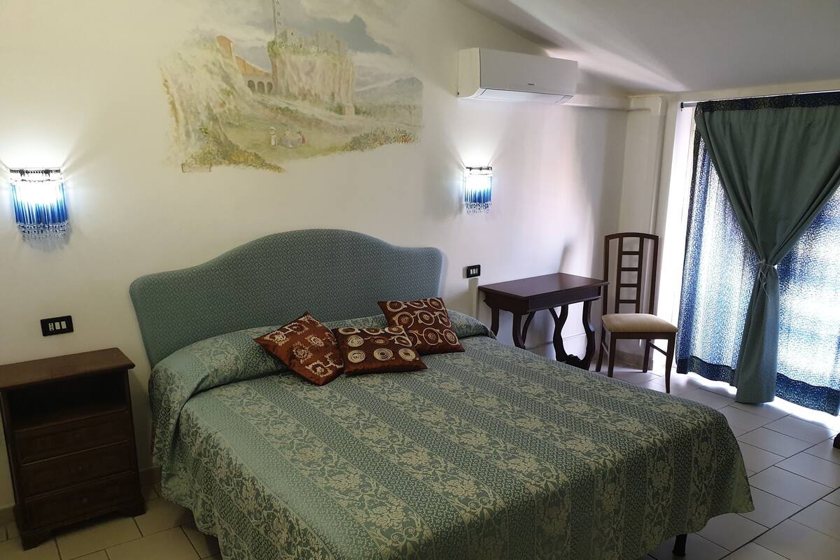 A bedroom with a large bed, a nightstand, a small table, a chair, and a wall-mounted air conditioner.