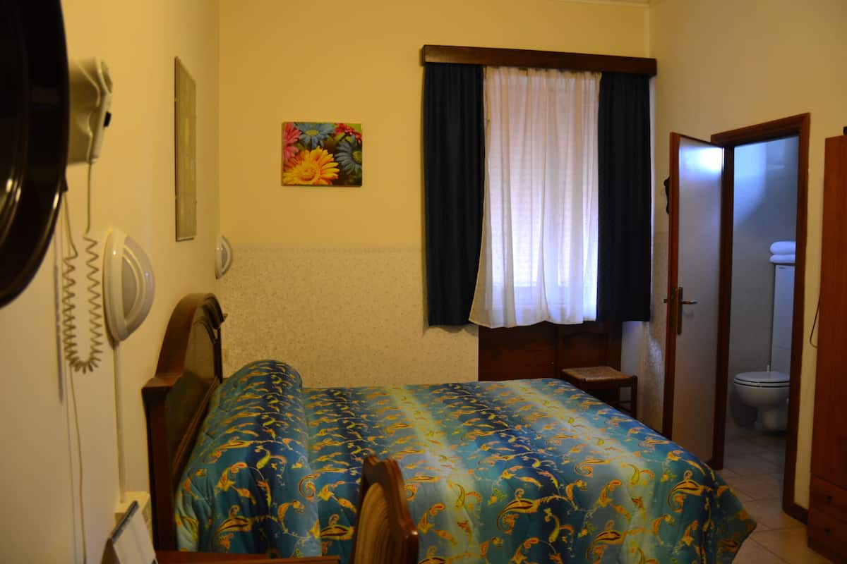 A bedroom with a bed, a bedside table, a chair, a window with curtains, a painting on the wall, and a bathroom visible through an open door.