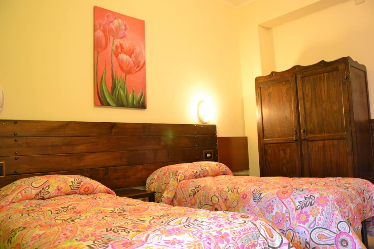 A hotel room with a wooden headboard, two beds with floral bedding, a nightstand, a wardrobe, and a wall-mounted light.