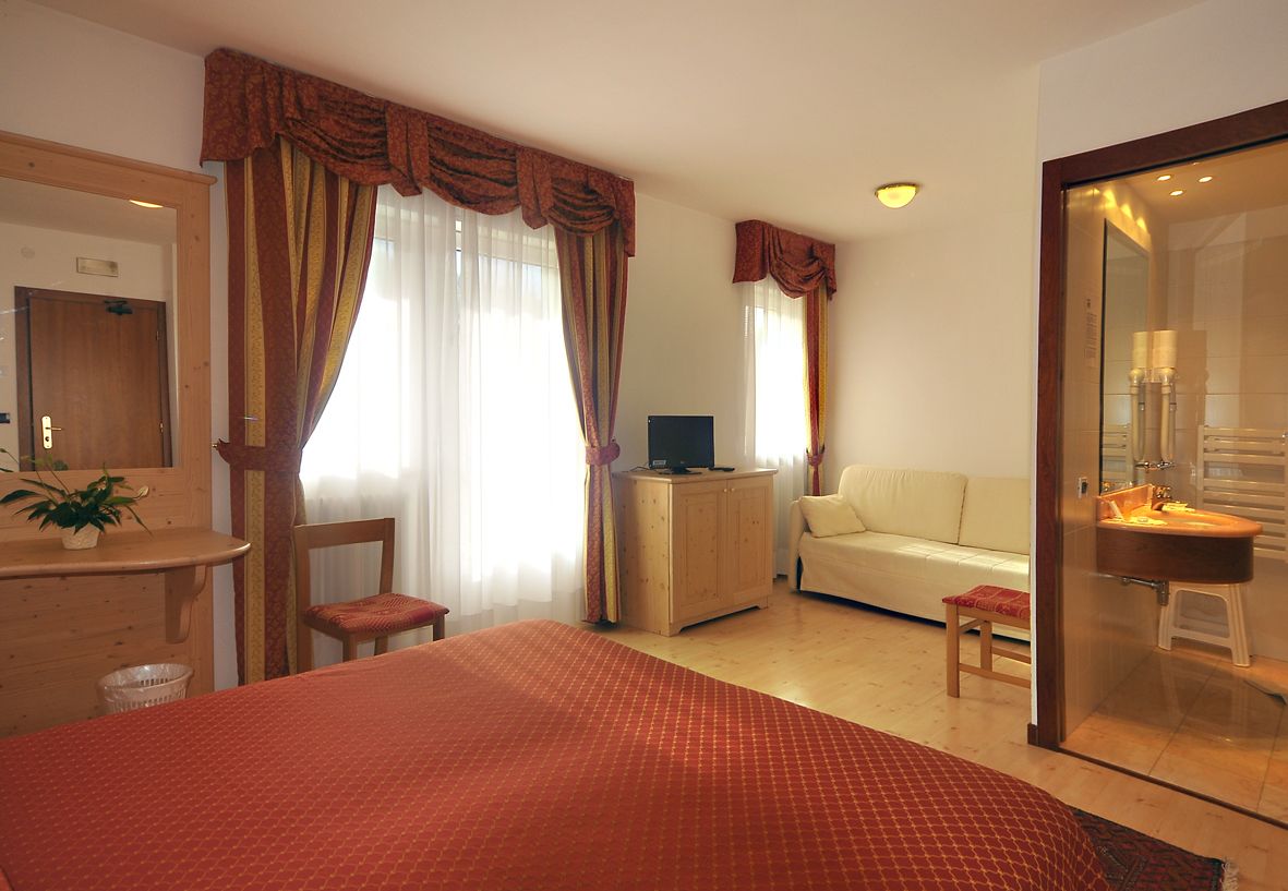 A hotel room with a bed, a desk, a chair, a sofa, a television, a small stool, and a bathroom with a sink and mirror.