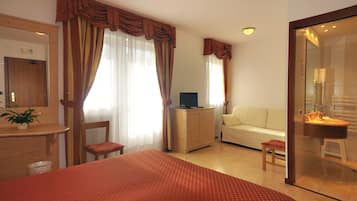 A hotel room with a bed, a desk, a chair, a sofa, a television, a small stool, and a bathroom with a sink and mirror.