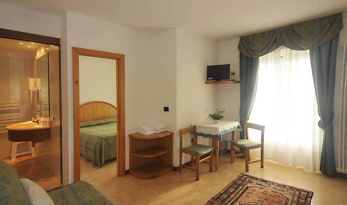 A compact hotel room with a bed, a small desk with a chair, a TV mounted on the wall, a window with curtains, and a bathroom with a sink and mirror.