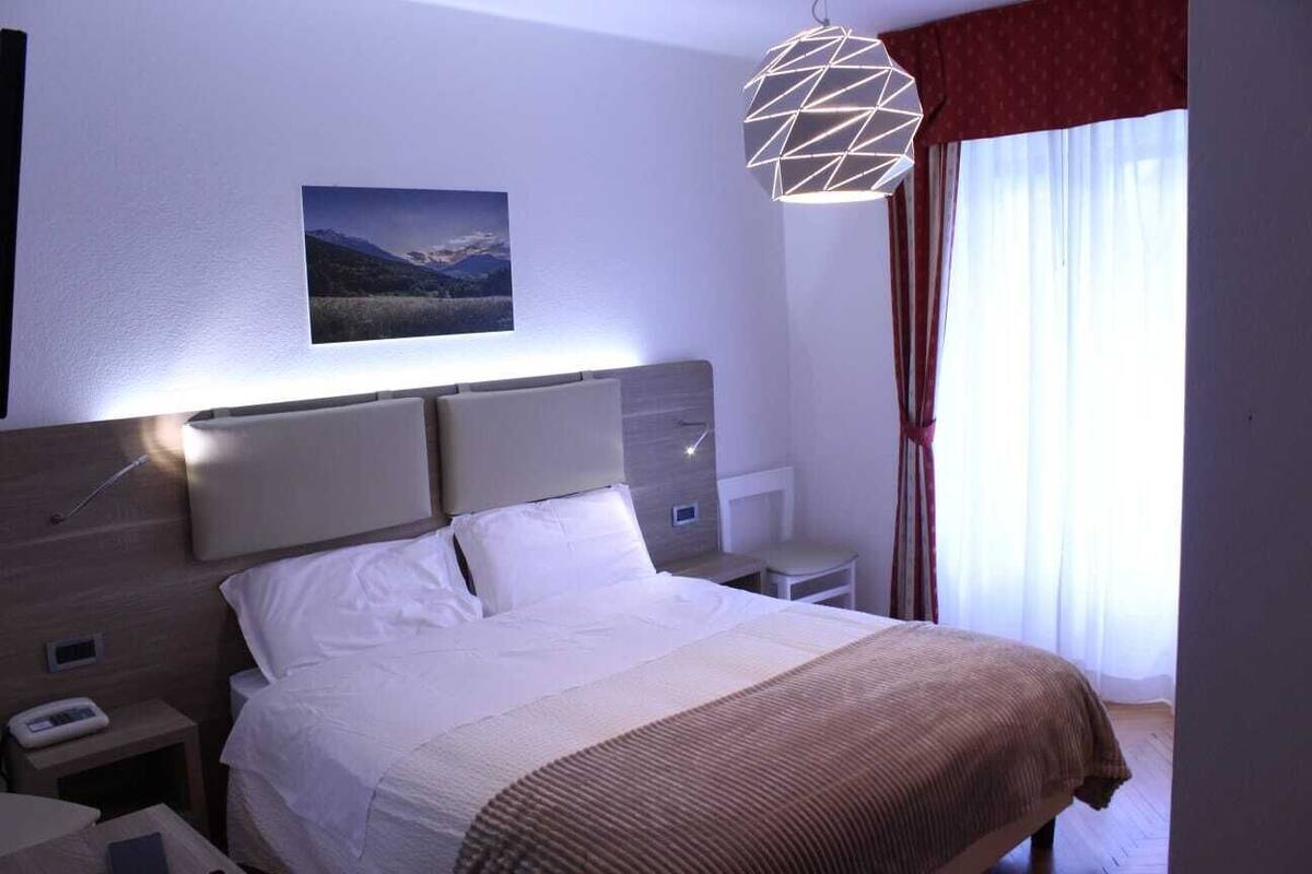 A hotel room with a large bed, a bedside table, a nightstand, a wall-mounted lamp, a picture, and a window with curtains.