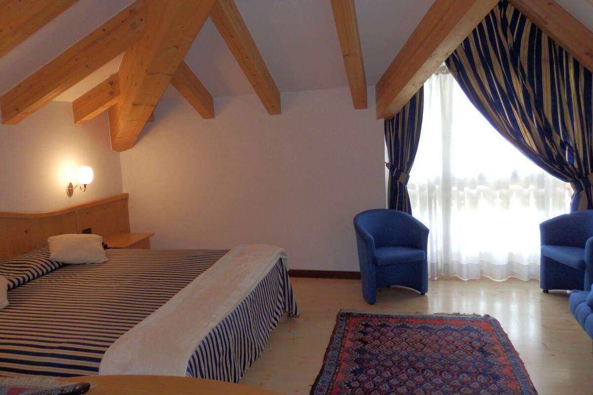A bedroom with a bed, two blue chairs, a patterned rug, wooden beams, and a window with curtains.