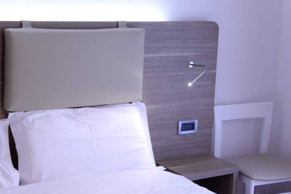 A hotel room with a bed, a bedside table, a chair, and a wall-mounted control panel.