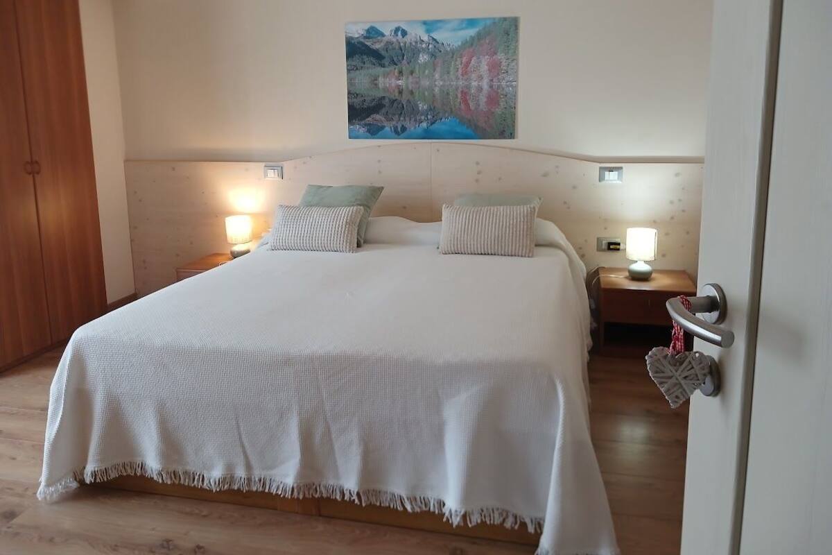 A bedroom with a large bed, two bedside lamps, a painting on the wall, and a wooden wardrobe.