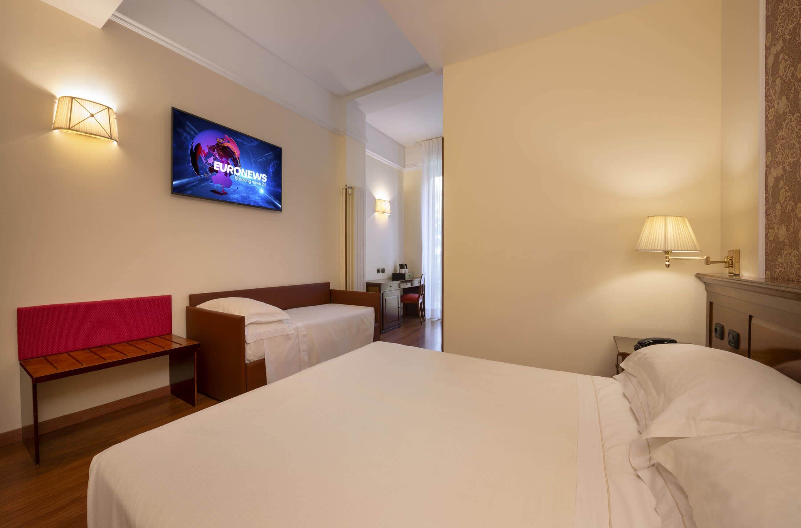 A hotel room with two beds, a desk, a chair, and a television displaying 'EURONEWS'.