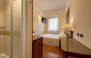 Una habitación de hotel con una cama, un televisor montado en la pared, una mesita de noche y una ventana con cortinas.