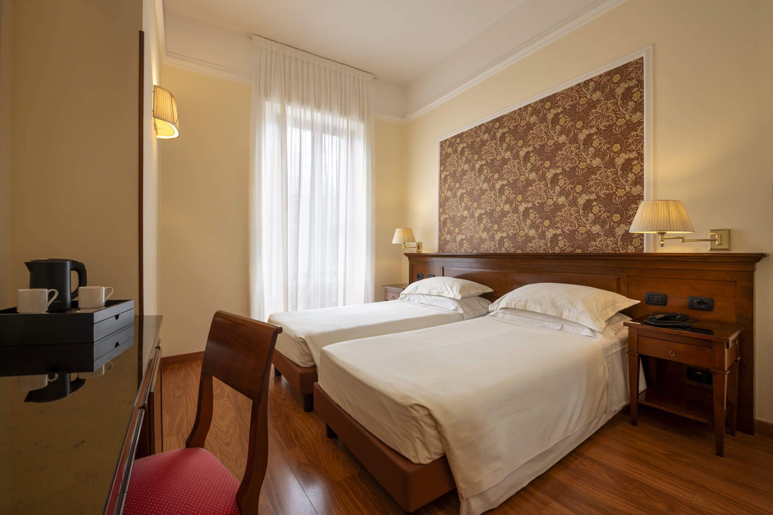 Camera d'albergo con due letti, un comodino in legno, una scrivania con lampada, una sedia e un grande muro con motivi floreali.