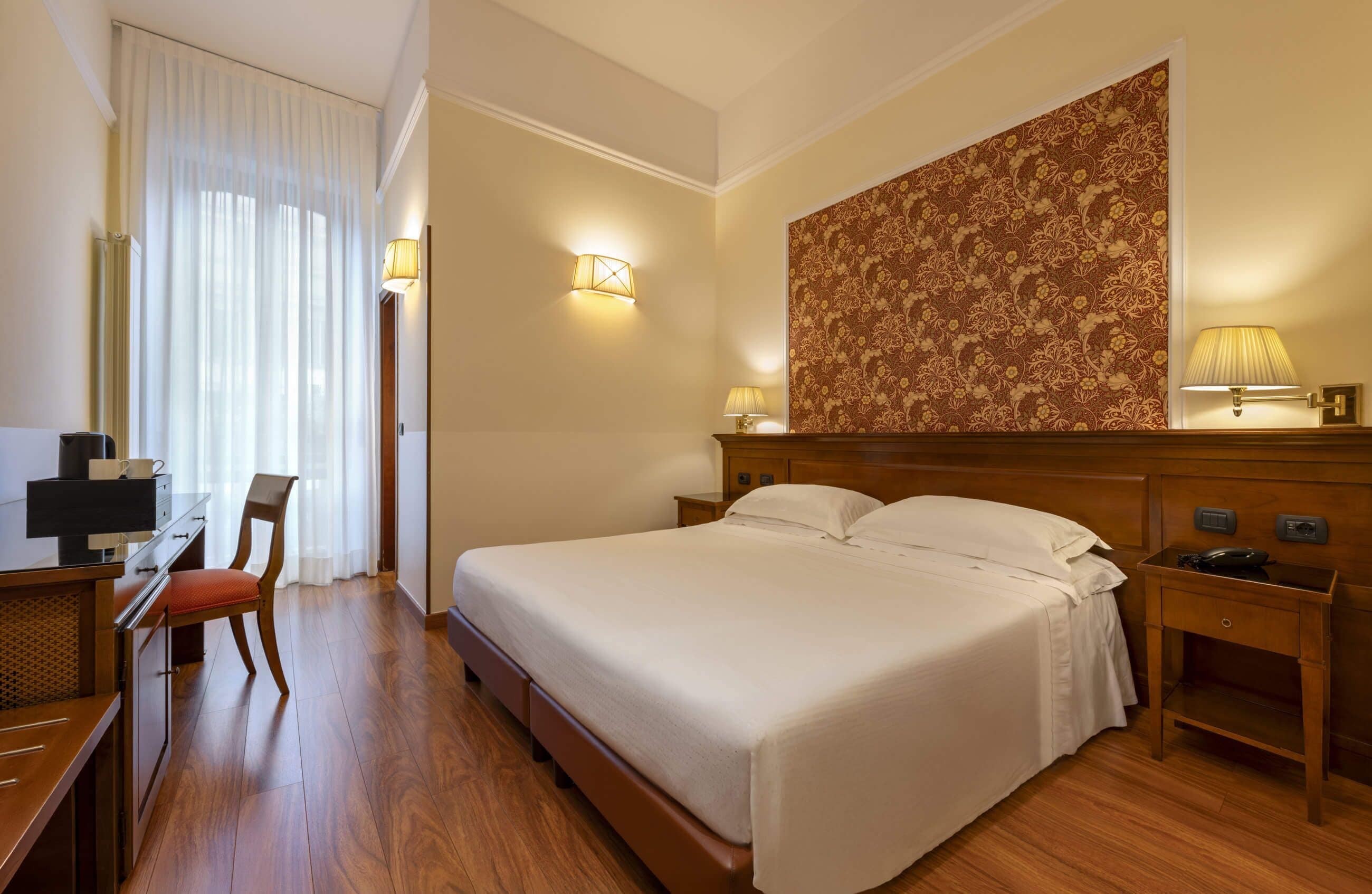 Ein Hotelzimmer mit einem großen Bett, einem Schreibtisch mit Stuhl, einem Kleiderschrank und einer Wand mit floralem Tapetenmuster.