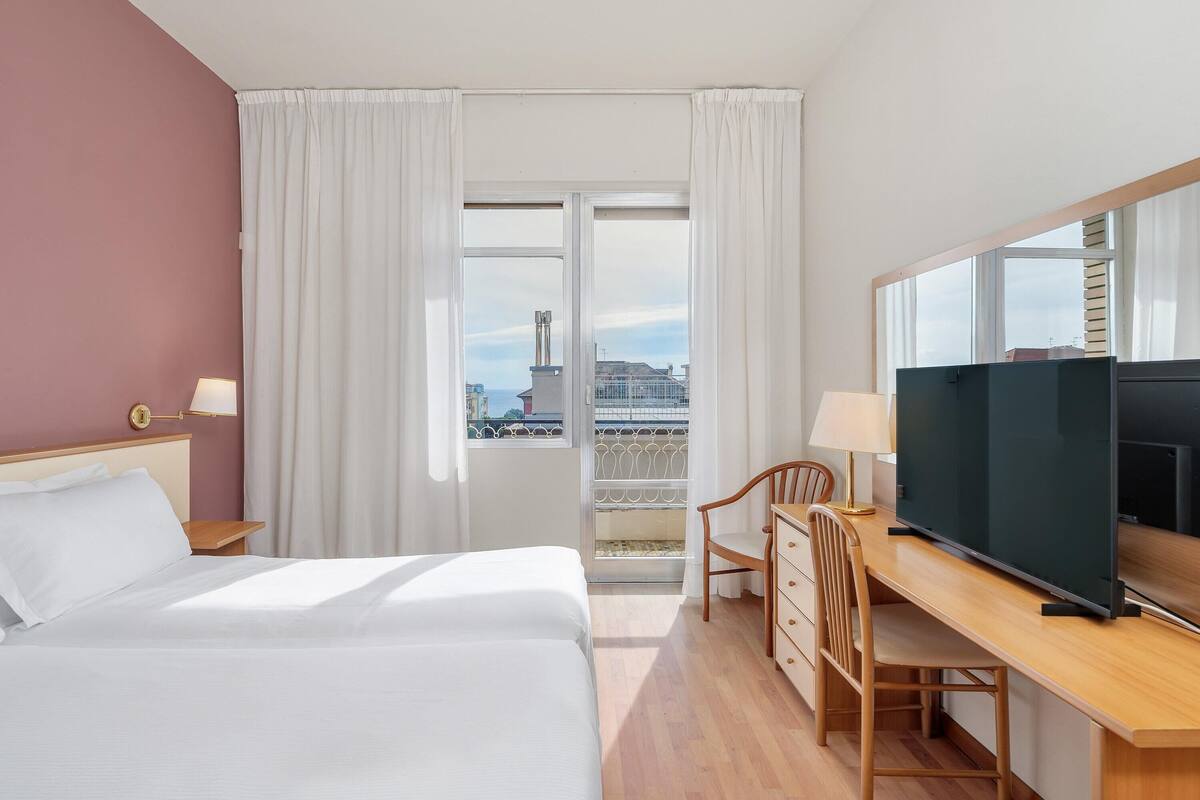 Une chambre d’hôtel avec un lit, un bureau avec une télévision, une chaise et une fenêtre avec des rideaux.