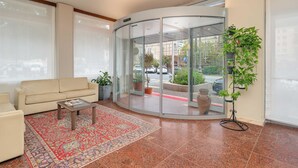 Vestíbulo de hotel con un sofá beige, una mesa de centro, un tapete estampado y una puerta de vidrio que da al exterior.