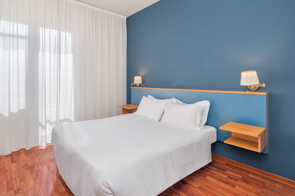 Une chambre à coucher avec un lit, du linge de lit blanc, deux tables de chevet avec des lampes, une petite table en bois et un mur d’accent bleu.