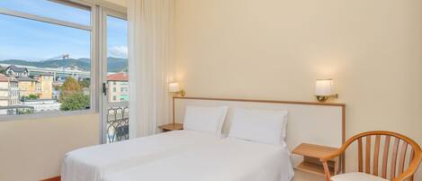 A hotel room with a large bed, wooden floor, two bedside lamps, a wooden chair, and a window with a view of buildings and mountains.