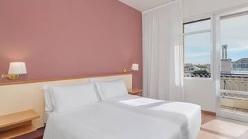 Une chambre d’hôtel avec un lit, un banc en bois, deux lampes fixées au mur et une grande fenêtre donnant sur la ville.