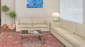 Un salón con un sofá beige, una mesa de centro con revistas, una alfombra estampada, un cuadro en la pared y una lámpara sobre una mesita auxiliar.
