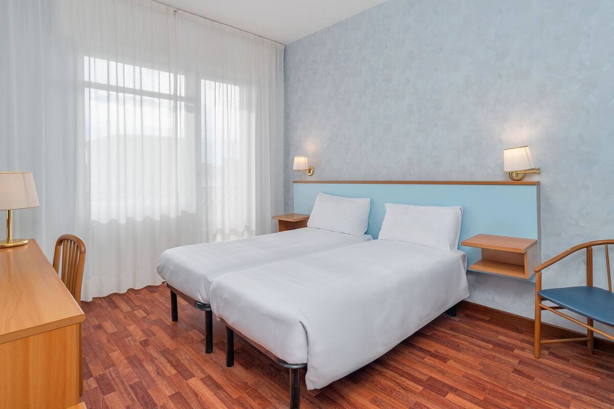 Une chambre d’hôtel avec un grand lit, deux tables de chevet, un bureau avec une chaise et une fenêtre avec des rideaux légers.
