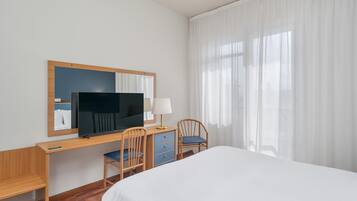 A hotel room with a bed, a desk with a TV, a chair, a lamp, and a window with curtains.