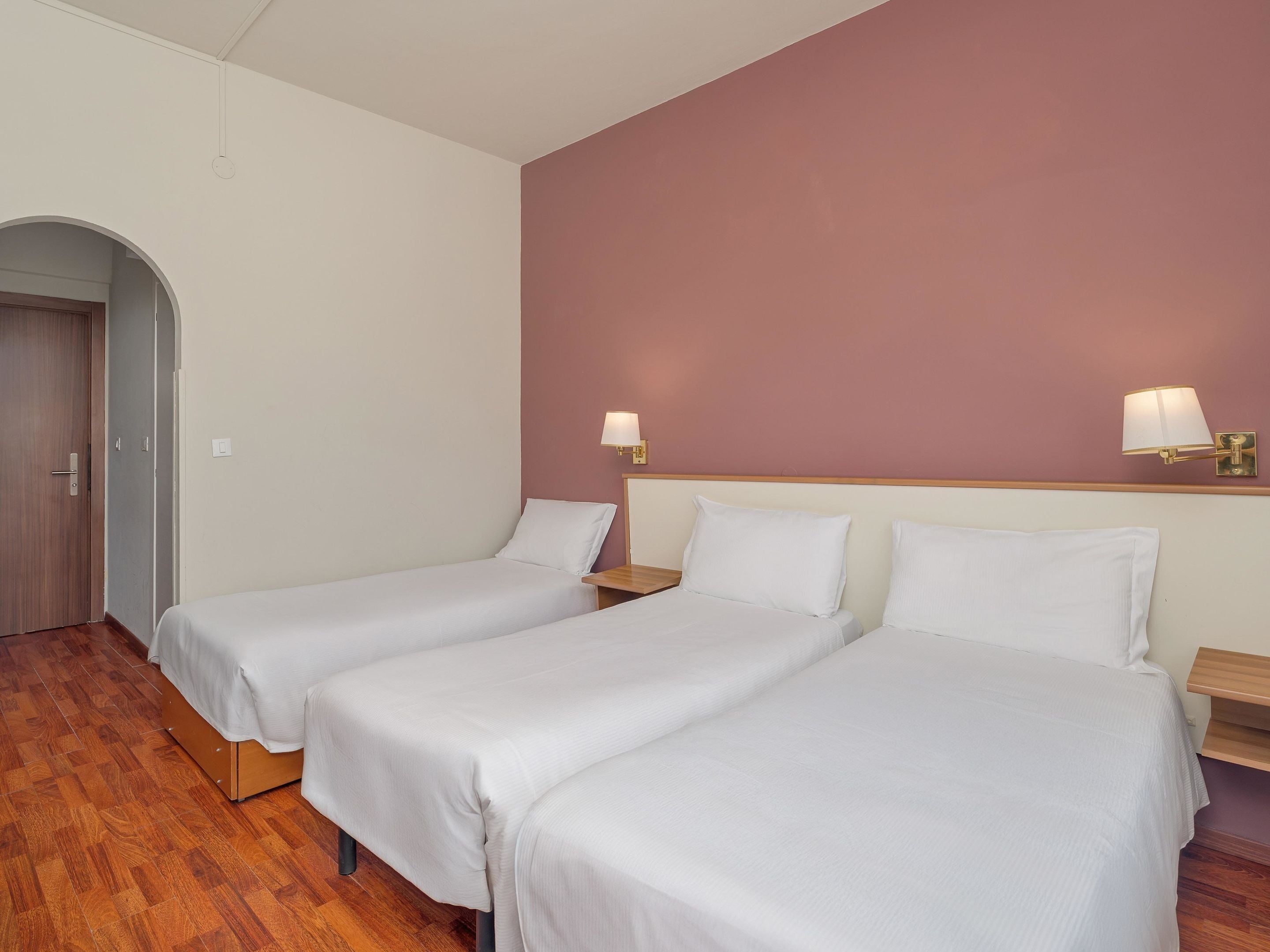 A hotel room with two single beds, wooden headboards, a wooden floor, and a pink accent wall.